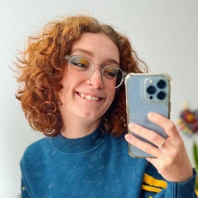 @AshleyRoboto on Twitter smiling at the camera, wearing a blue sweater, and holding her phone to take a mirror selfie with a blurry light colored background